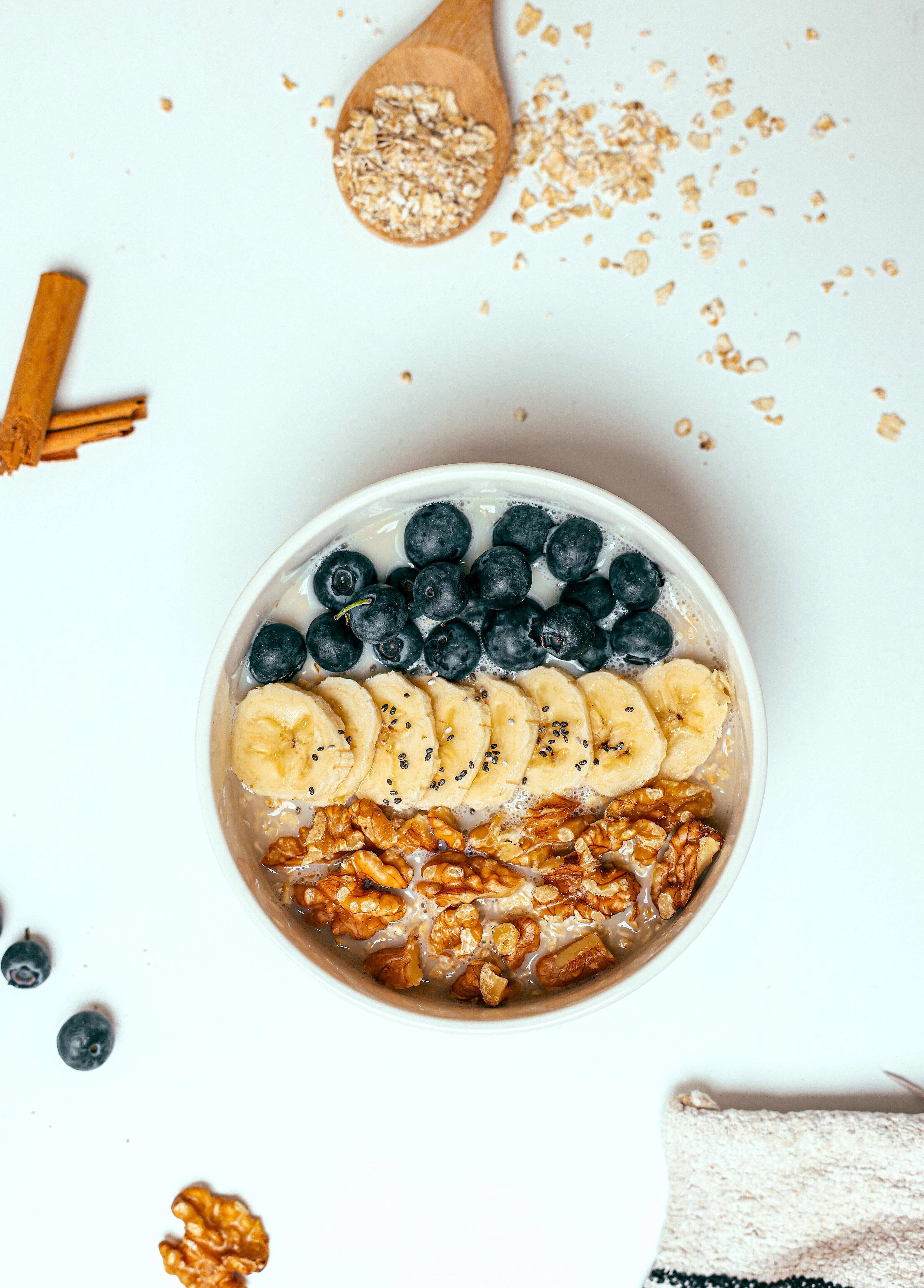 A Smoothie Bowl with Banana, Blueberry, Nut Topping on a White Surface surrounded by gronala, a spoon, cinnamon, and a towel.