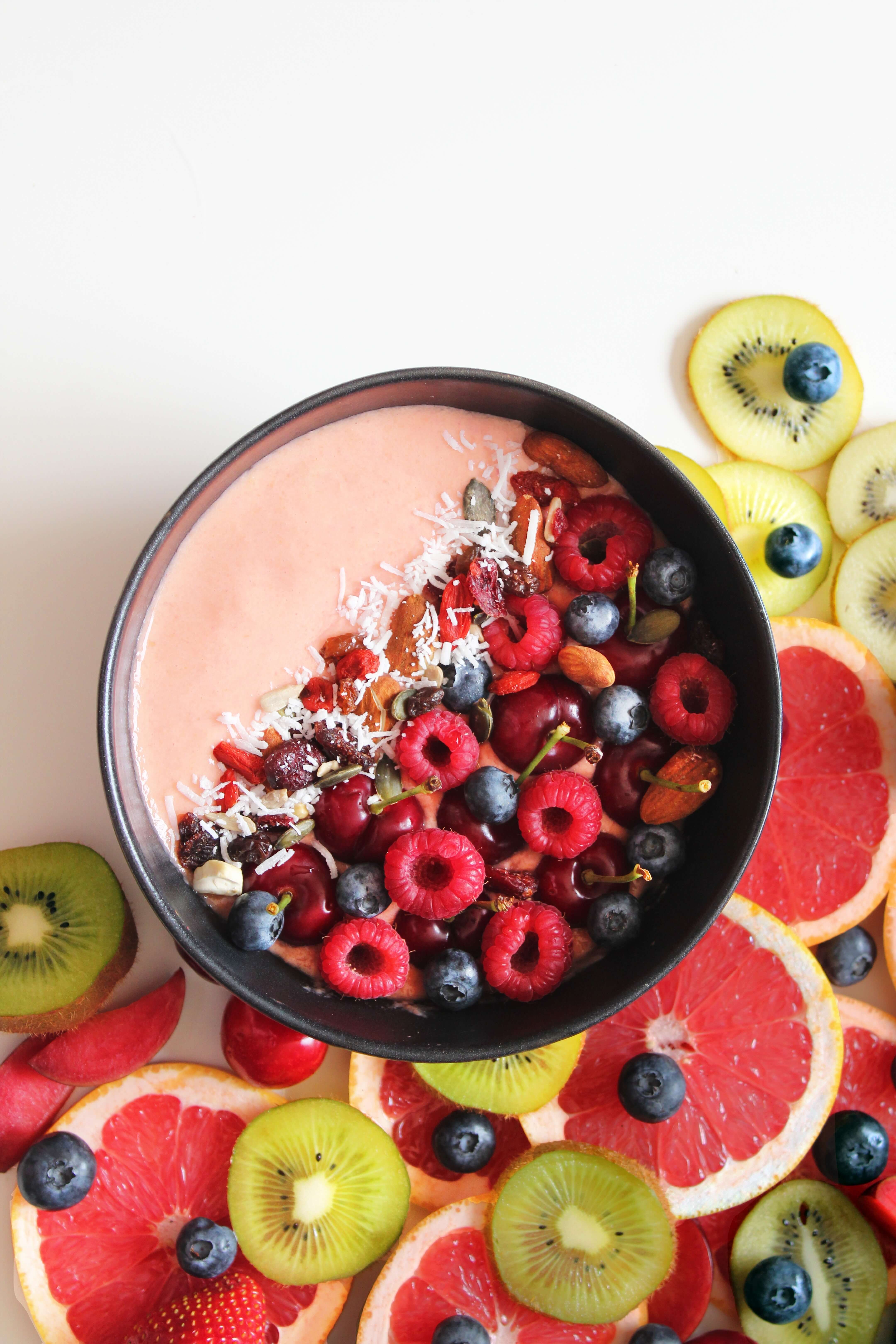 A Smoothie Bowl with Fresh Fruits Topping and Fruit around its right corner on a White Surface.