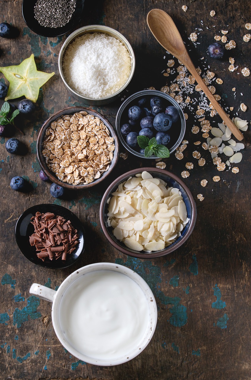 Ingredients for making smoothie for healthy breakfast. Bowls of yogurt, blueberries, granola, almond chia seeds, coconut, milk, chocolate, mint, carambola over dark wooden background 